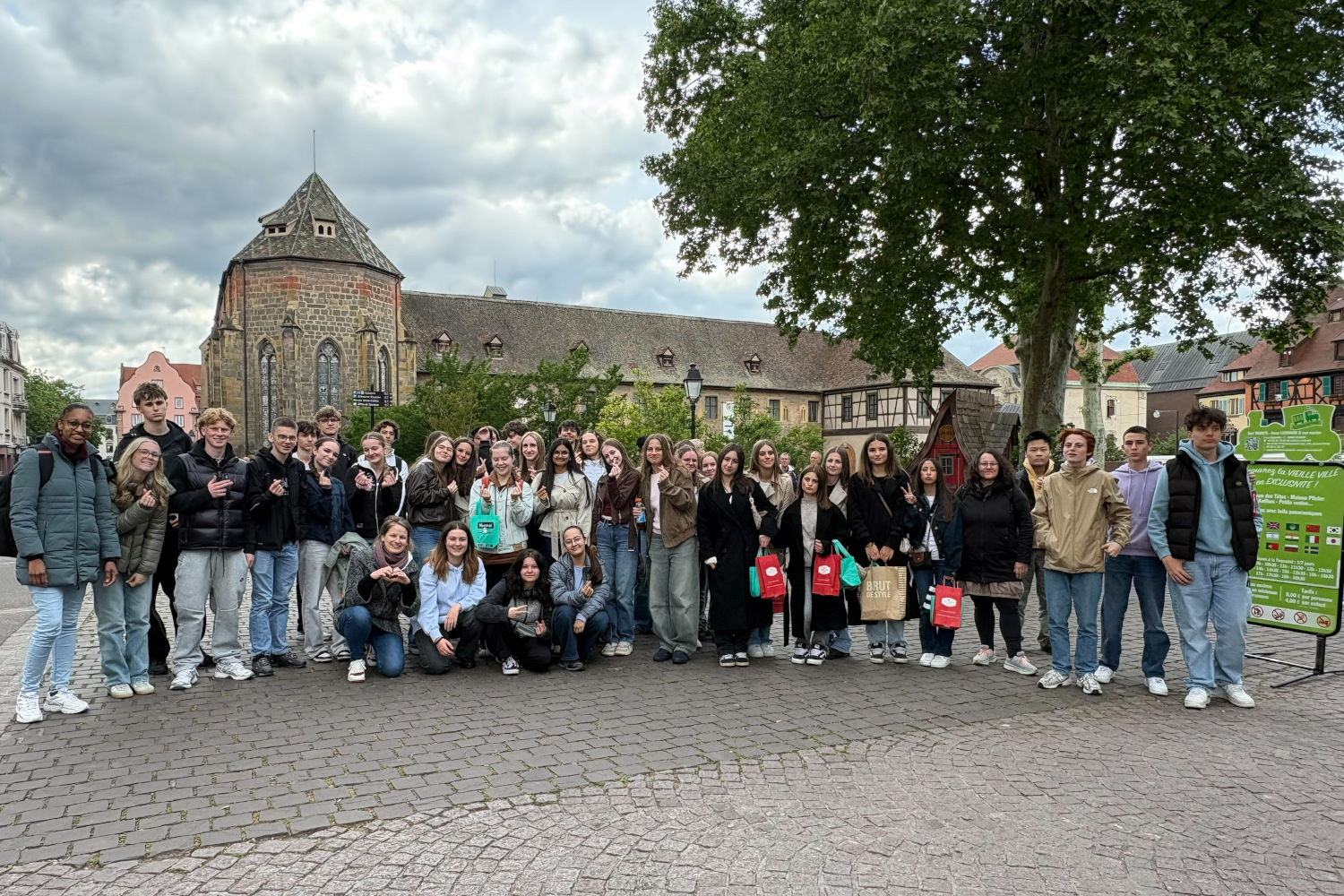 Abschlussfoto einer großen Gruppe von Jugendlichen der Bertha-von-Suttner-Schule mit ihren Lehrkräften vor der Heimreise. Sie stehen auf dem gepflasterten Platz an der Rue Kléber vor der historischen Kulisse des Unterlinden-Museums.