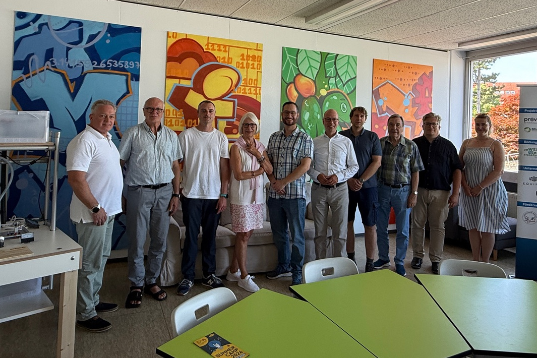 Gruppenfoto im MINT-Zentrum Nidderau vor bunten Graffiti-Kunstwerken an der Wand. Zehn lächelnde Personen stehen nebeneinander in einem hellen Raum, darunter Dr. h.c. Beate Heraeus (Mitte, mit weißer Brille und rosa-weißem Outfit). Im Vordergrund sind hellgrüne Tische zu sehen, auf einem liegt ein Flyer mit der Aufschrift „Summer Camp 2026“. Rechts im Hintergrund steht ein Roll-up-Banner mit Logos der Partnerorganisationen.