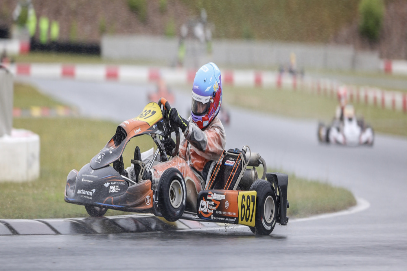 Ein junger Kart-Rennfahrer mit blauem Helm und orangefarbenem Rennanzug steuert ein Go-Kart mit der Startnummer 687 der Marke Dörr Motorsport dynamisch durch eine Kurve auf einer nassen Rennstrecke. Das Kart neigt sich in der Kurve leicht zur Seite. Im verschwommenen Hintergrund ist die Rennstrecke mit Curbs und einem weiteren Kart zu sehen.