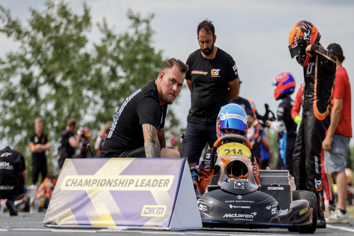 Ein junger Kartfahrer mit blau-orangefarbenem Helm sitzt in seinem Rennkart mit der Startnummer 214. Neben dem Kart kniet ein Mechaniker in einem schwarzen T-Shirt von Dörr Motorsport. Im Vordergrund steht ein Aufsteller der DKM mit der Aufschrift „Championship Leader“.