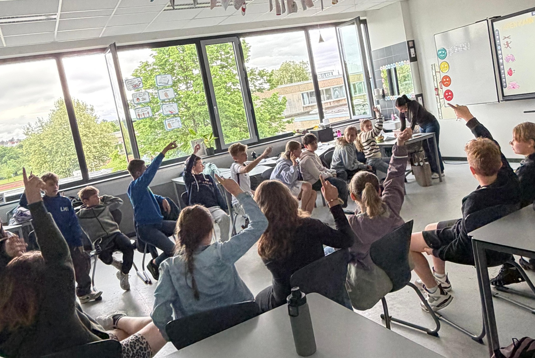 Schülerinnen und Schüler der 5. Klasse sitzen in einem Stuhlkreis in ihrem hellen Klassenzimmer an der Bertha-von-Suttner-Schule. Sie nehmen aktiv an dem Suchtpräventions-Workshop „Rauchzeichen“ teil, während viele von ihnen motiviert die Hände heben, um sich zu beteiligen. Im Hintergrund ist eine Lehrkraft am Whiteboard zu sehen, auf dem Symbole zur Suchtprävention abgebildet sind. Große Fensterfronten geben den Blick ins Grüne frei und sorgen für eine freundliche Lernatmosphäre.